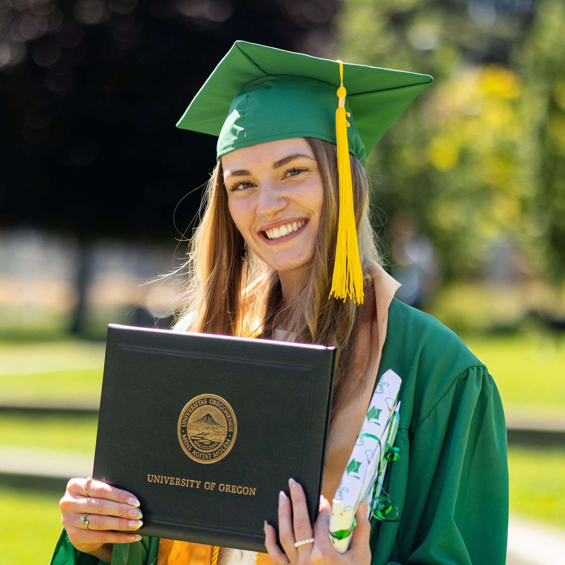 A smiling graduate in a green cap and gown holds a University of Oregon diploma cover and a decorated diploma, standing outdoors on a sunny day.