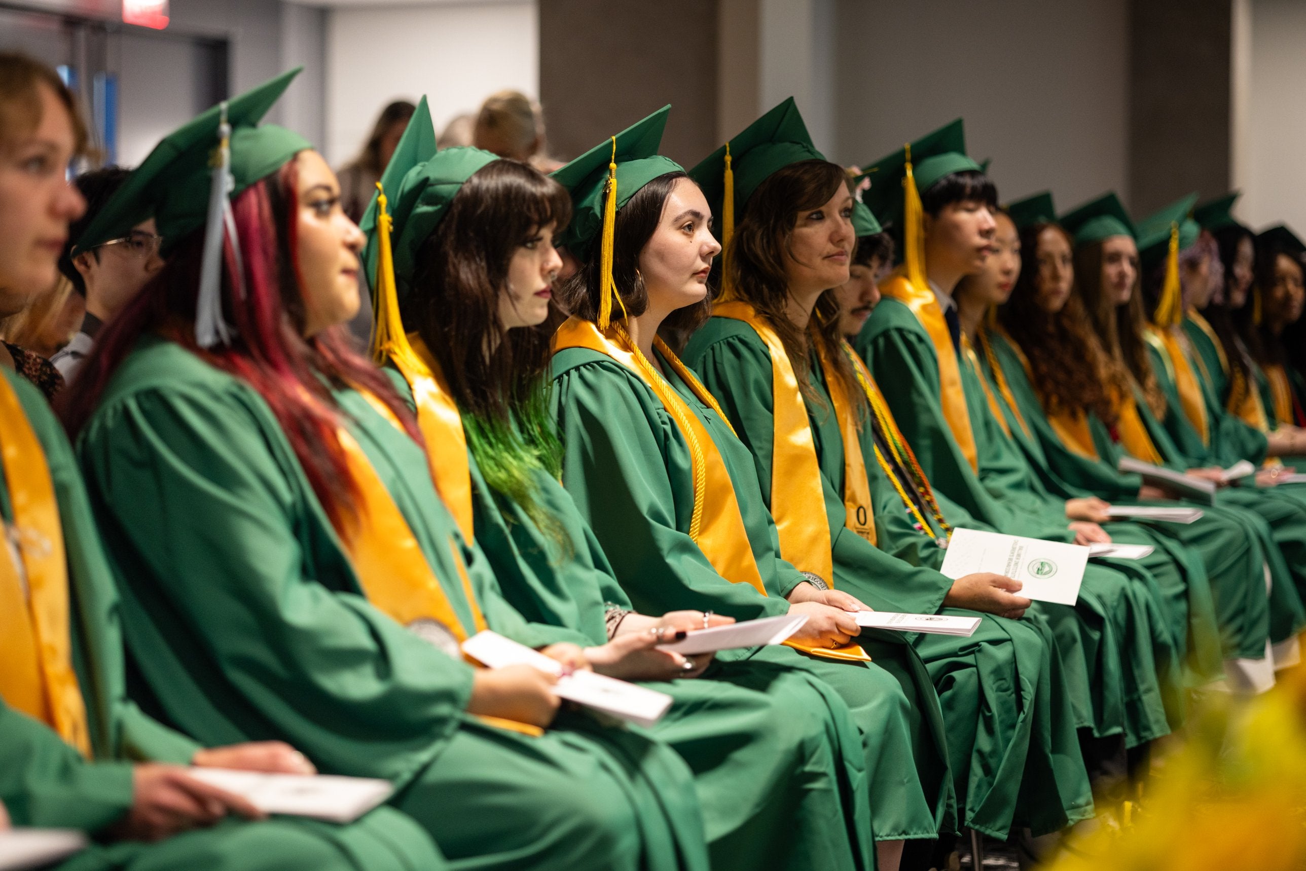 Ballmer Institute graduates in green caps and gowns with gold stoles sit in a row during commencement