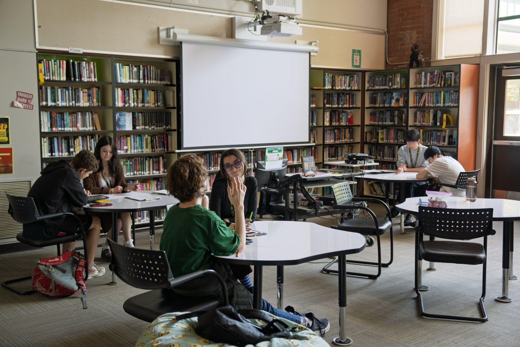Students work and talk at tables in a school library while a child behavioral health student meets with a young person