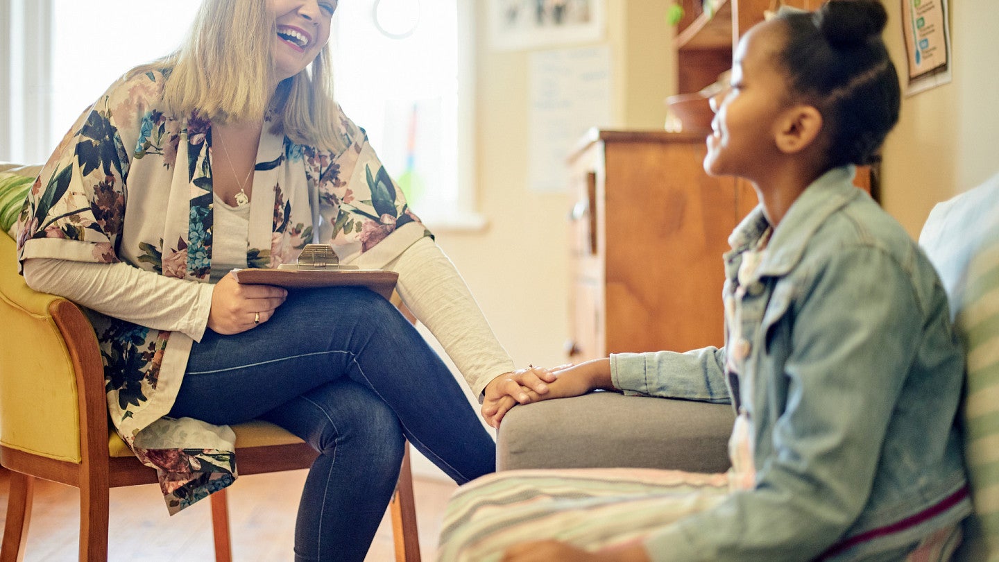 A smiling counselor holding a clipboard sits across from a young girl during a warm, relaxed one-on-one session indoors.