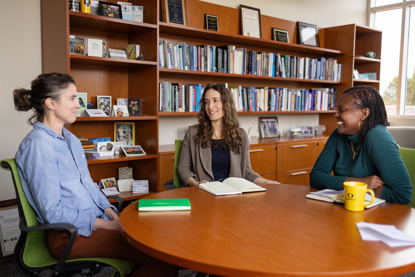 three women talking in an office setting with books behind them