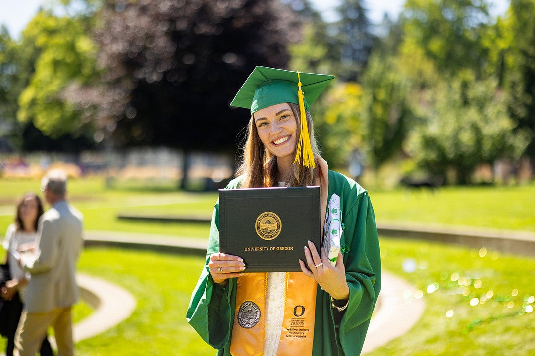 A graduate wearing a green cap and gown holds a University of Oregon diploma. In the background, people are gathered outdoors on a sunny day with trees and grass surrounding them.