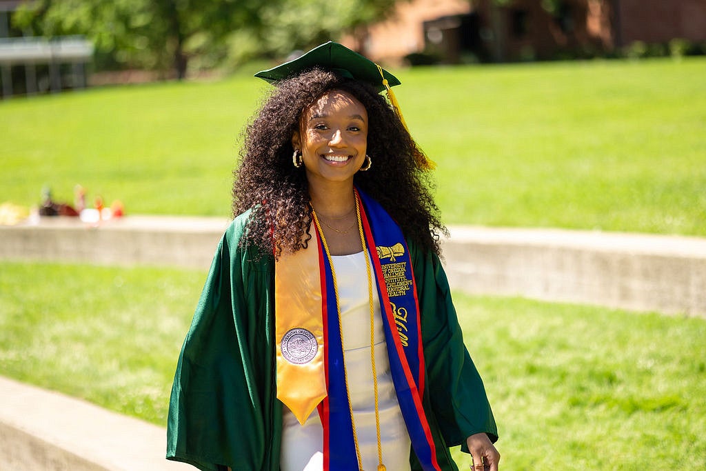 A smiling graduate in a green cap and gown walks outdoors on a sunny day, wearing multiple honor stoles including a gold University of Oregon stole and a blue Ballmer Institute for Children's Behavioral Health stole.