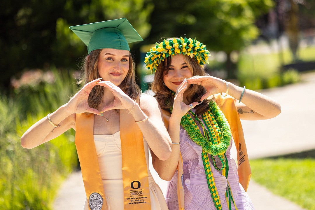Two people are posing outdoors, wearing graduation stoles with the University of Oregon logo. One wears a green cap and gown, and the other wears a purple outfit with a decorative floral headpiece and leis. Both are making the shape of the letter "O" with their hands. The background features greenery and a walkway.