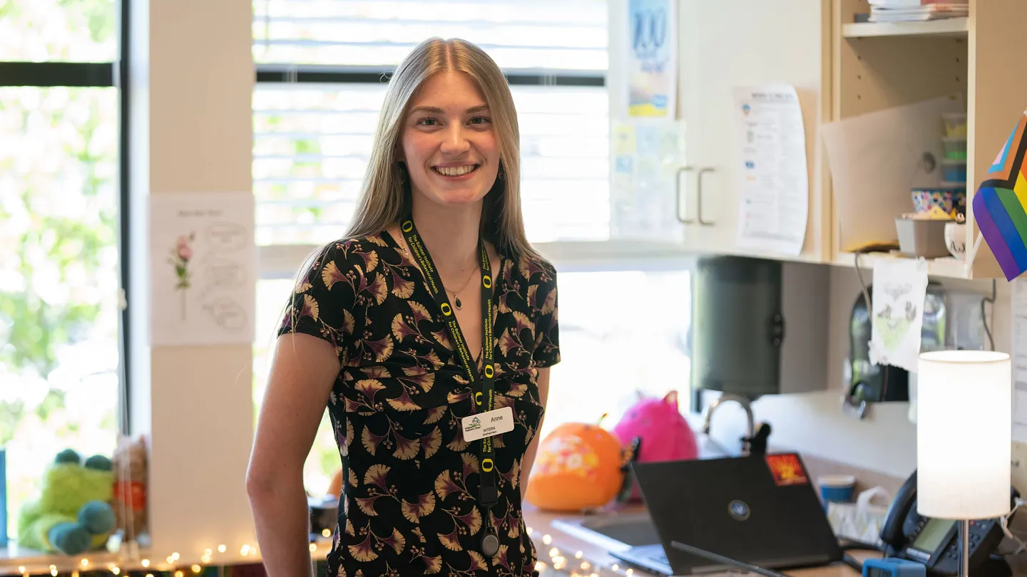 Anne Libby, a Ballmer Institute intern, smiles while standing at a clinic reception desk wearing a UO lanyard and name badge.