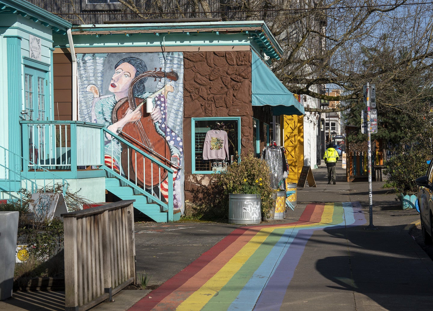A colorful sidewalk painted like a rainbow runs past a building with a mural of a musician playing cello; a person in a yellow jacket walks in the distance.