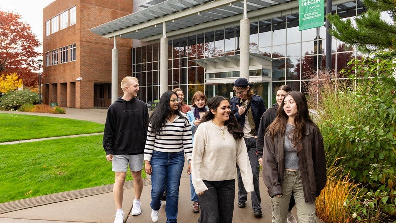 A group of students walk together on a sidewalk outside a modern campus building with large windows and a green lawn.