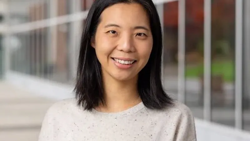 Headshot of a woman with straight black hair smiling outdoors in front of a modern campus building