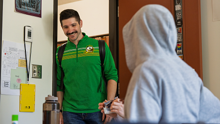 A person wearing a green University of Oregon shirt and backpack smiles while entering a classroom and greeting a student in a gray hoodie. Posters and stickers promoting inclusion and anti-violence messages are visible on the door and wall.
