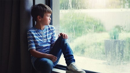 A young boy in a striped shirt and jeans sits on a window ledge, looking out at a rainy outdoor scene.
