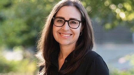 Smiling woman with glasses and shoulder length brown hair standing in front of greenery