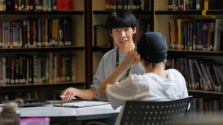 Two people sit across from each other at a table in a library, with bookshelves in the background