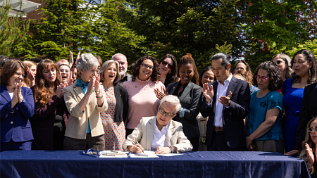 Governor Tina Kotek signs behavioral health legislation at an outdoor ceremony at the Ballmer Institute, surrounded by applauding legislators and advocates.