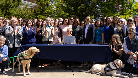 Governor Tina Kotek holds up the signed behavioral health legislation surrounded by a large group of legislators, advocates, and providers — plus two service dogs — outside the Ballmer Institute.