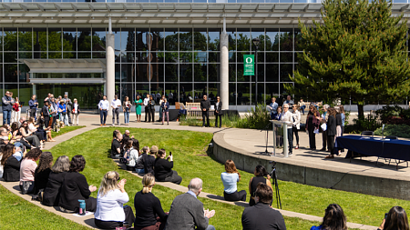 Attendees gather on the lawn outside the University of Oregon's Ballmer Institute for the outdoor bill signing ceremony, where speakers address the crowd from a podium in front of the building.