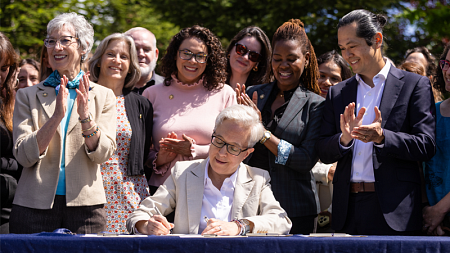 Governor Tina Kotek signs behavioral health workforce legislation at the University of Oregon's Ballmer Institute, surrounded by applauding legislators, advocates, and behavioral health providers at an outdoor bill signing ceremony.