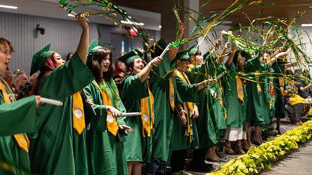 A row of graduates in green caps and gowns cheer and wave green and gold tinsel streamers indoors at a commencement ceremony. They hold diplomas and wear gold honor stoles, with yellow flower arrangements lining the floor in front of them and an audience visible in the background.