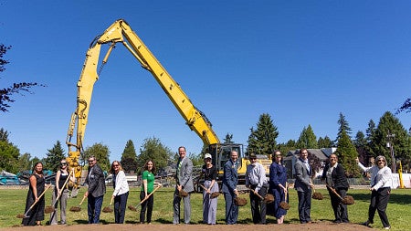 Thirteen people pose with shovels at a groundbreaking ceremony, with a large yellow excavator behind them under a clear blue sky.