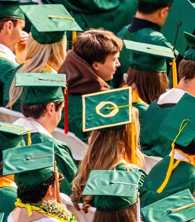 verhead view of graduates wearing green caps and gowns seated in rows at an outdoor commencement ceremony.