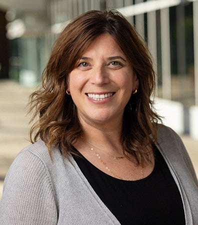 Headshot of a woman with brown wavy hair smiling outdoors in front of a modern campus building