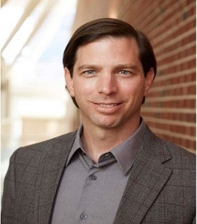 a man smiles for a portrait with a brick facade behind him