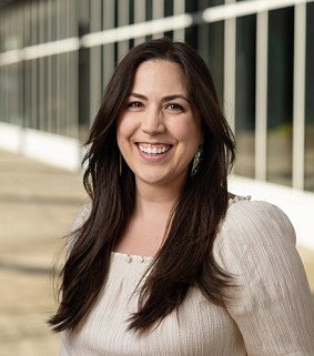 Smiling woman with long dark brown hair wearing a light beige blouse, standing in a sunlit walkway with glass windows in the background.