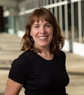 Smiling woman with shoulder-length light brown hair wearing a black short-sleeve top, standing in a sunlit walkway with glass windows in the background