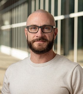 Man with a beard and glasses wearing a light beige sweater, standing in a bright outdoor walkway with glass windows in the background.