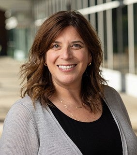 Smiling woman with shoulder-length brown hair wearing a black top and light gray cardigan, standing in a bright hallway with glass windows.