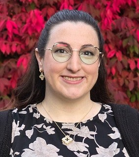 Smiling woman with dark pulled-back hair and glasses wearing a black and white patterned shirt with a black cardigan stands in front of a red background.