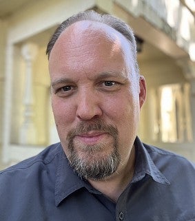 A bearded man wearing a blue button-down poses in front of a building