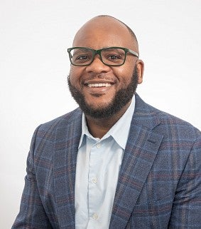A smiling man with a beard and glasses wearing a blue plaid blazer and white shirt poses in front of a white background.