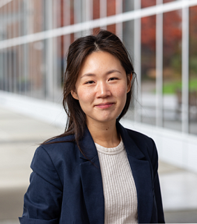 A smiling woman with dark hair wearing a white shirt and blue blazer poses in front of a wall of windows