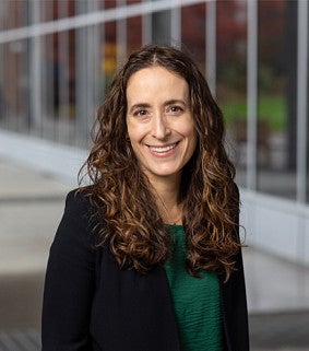 Smiling woman with long, curly hair wearing a black blazer over a green top, standing outdoors near a modern glass building.