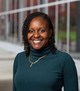 A woman wearing a dark green turtleneck and gold necklace, standing outdoors near a modern glass building.