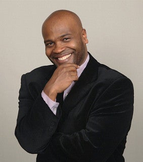 A smiling man wearing a black suit and striped shirt poses with his hand on his chin