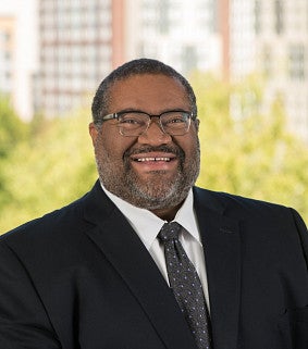 A man wearing glasses, a dark suit, and a tie smiles outdoors with green trees and tall buildings in the blurred background.