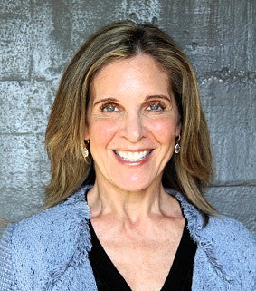A woman with long, light brown hair smiles at the camera. She is wearing a light blue jacket over a black top, standing in front of a gray textured wall.