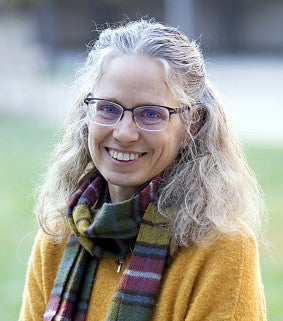 Smiling woman with long gray hair and glasses, wearing a yellow sweater and a green plaid scarf, standing outside with a blurred background of grass.