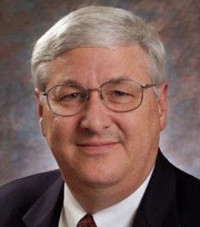 A man with short gray hair and glasses is wearing a dark suit, white shirt, and red tie, smiling slightly in a formal portrait with a blurred gray and brown background.