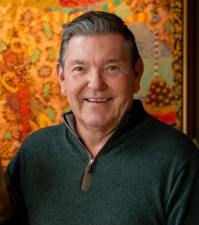 A smiling man with short gray hair wearing a dark green sweater, standing in front of a colorful, patterned background.