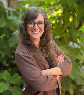 Emily Kroman smiling, with long brown hair and glasses, wearing a brown cardigan with her arms crossed, standing in front of green leafy foliage.
