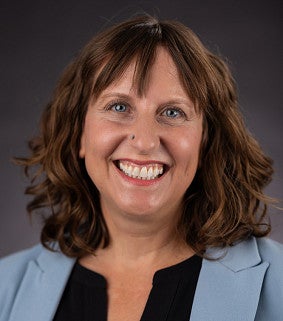 Headshot of Erika K. Coles smiling, with shoulder-length wavy brown hair and bangs, wearing a gray blazer over a black top, against a dark gray background.