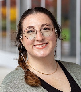 Han Padilla smiling, wearing glasses with a beaded eyeglass chain, a braid, and a gray cardigan over a black top