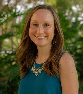 Kelsey Dickson, smiling, with long auburn hair, wearing a teal sleeveless top and statement necklace, outdoors with green foliage behind her.