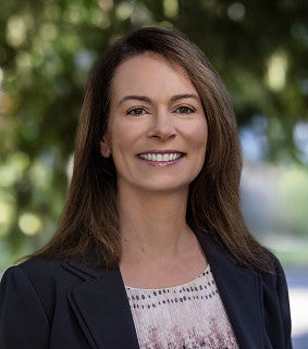 Karen Chaput smiling, with long brown hair, wearing a black blazer over a patterned blouse, outdoors in front of blurred greenery.