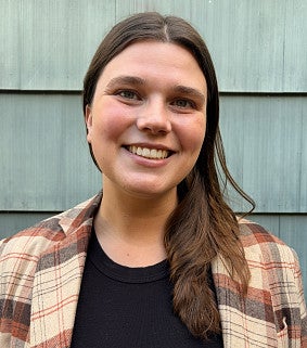 Madeleine Allen smiling, with long brown hair and a plaid flannel jacket over a black top, in front of gray wood siding.