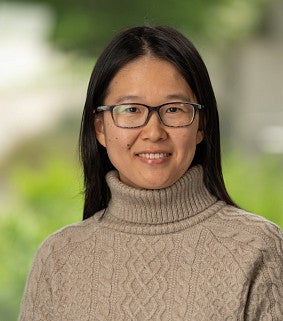 Yu Zhang smiling, with long black hair and glasses, wearing a tan cable-knit turtleneck sweater against a blurred green outdoor background.