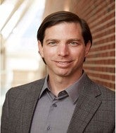 a man smiles for a portrait with a brick facade behind him