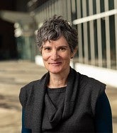 Smiling woman with short hair wearing a gray sweater top, standing in a bright hallway with glass windows.